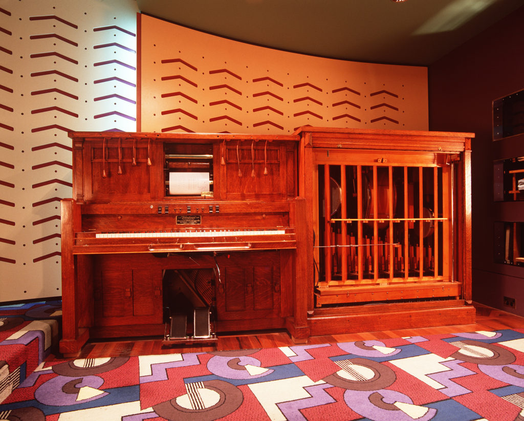 An upright piano attached to a large wooden case, of the same height, containing a variety of musical instruments. At the base of the piano are two large pedals, and above the keys is a scroll of music with four leather pulleys on either side.